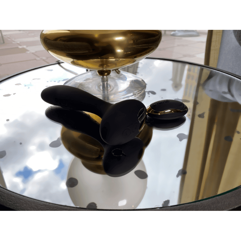 Gaea vibrating black dildo on a mirrored table beside a decorative bowl, showcasing its sleek design and remote control.